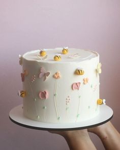 a hand holding a white cake with flowers and bees on the frosting, in front of a pink background