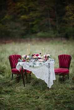 two red velvet chairs and a table with flowers on it in the middle of a field