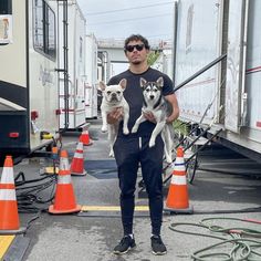 a man holding two dogs in his arms while standing next to some cones and trucks