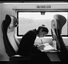 a person writing on a paper while sitting in a seat next to a window and looking out the window