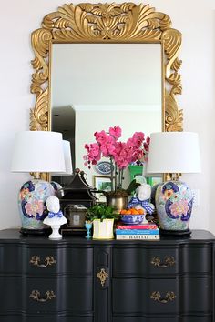 a black dresser topped with lots of flowers and vases next to a large mirror