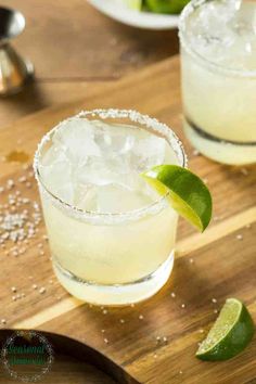 two margaritas sitting on top of a wooden cutting board next to lime wedges