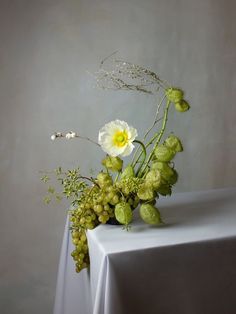 a vase with flowers and greenery on a white tableclothed table cloth in front of a gray wall