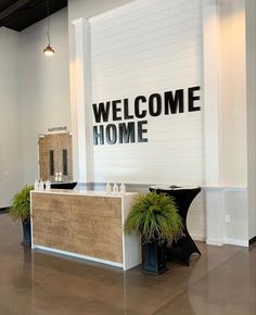 a welcome sign is on the wall behind a reception desk with two potted plants