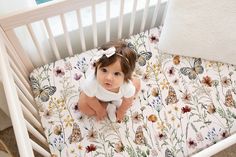 a baby sitting in a white crib next to a pillow with butterflies on it