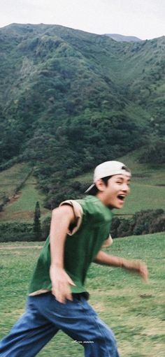 a young man running across a lush green field next to a mountain covered in trees