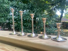 four silver candlesticks lined up on a wooden table outside in the sun with trees behind them