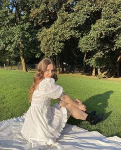 a woman sitting on top of a white blanket in a field next to trees and grass