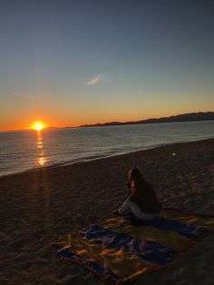 a woman sitting on top of a beach next to the ocean under a sun set