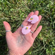 a hand holding two pink flowers on it's fingers in the grass with one flower sticking out of its middle