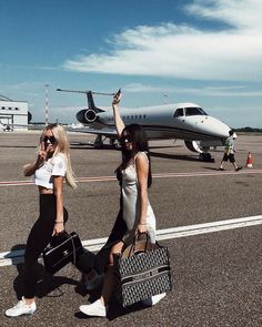 two beautiful women walking across an airport tarmac with luggage in hand and a plane behind them