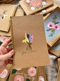 a hand holding a spiral notebook with flowers on it and the cover is lined in brown paper