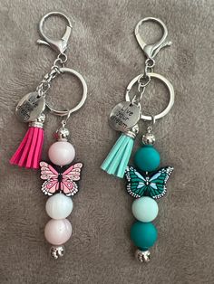 two key chains with charms attached to them on a blanket, one has a butterfly and the other has a tassel