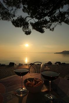 two glasses of wine sitting on top of a wooden table next to the ocean at sunset