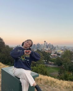 a woman sitting on top of a green box next to a lush green field and tall buildings