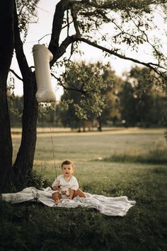 a baby sitting on a blanket under a tree with an object hanging from it's side