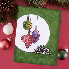 a christmas card with ornaments hanging from it's side on a pink surface next to pine cones