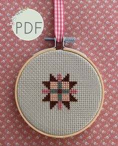 a cross - stitch pattern is hanging from a hoop on a pink background with a red and white checkered ribbon