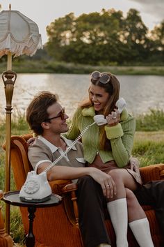 a man and woman sitting on an old fashioned chair talking on the phone, with a lake in the background