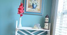 a white shelf with flowers and a vase on it next to a window in a blue room