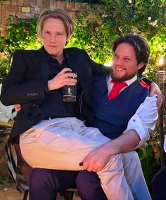 two men sitting next to each other in front of some trees and bushes, one holding a drink