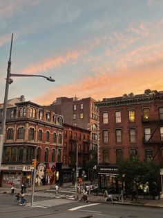 people are crossing the street in front of some buildings at sunset or sunrise, with pink clouds overhead