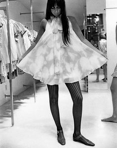 black and white photograph of a woman in a dress with tights on, standing next to clothing racks