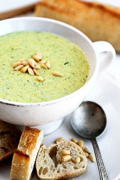 a white plate topped with a bowl of broccoli soup next to slices of bread