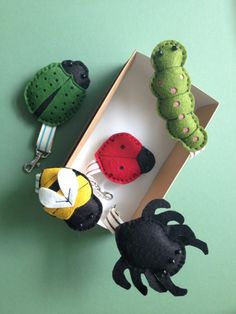 three stuffed animals are in a box on a green surface, one is holding a fork and the other has a caterpillar