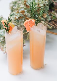 two tall glasses filled with orange juice and garnished with rosemary, sitting on a white surface