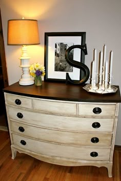 a white dresser with candles and pictures on it next to a framed letter, flowers and a lamp