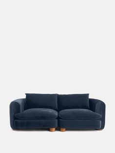 a blue velvet couch with wooden legs and pillows on the back, in front of a white wall