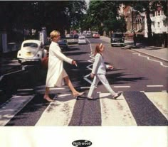 two women are crossing the street in front of an old white car and some buildings