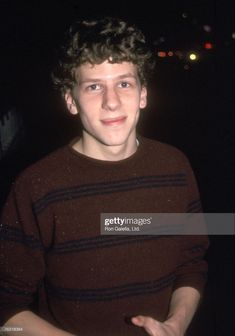 a young man standing in front of a dark background wearing a striped sweater and holding his hands out