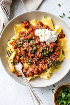 a white bowl filled with pasta and meat sauce on top of a table next to a fork