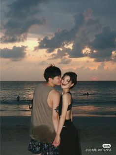 a man and woman kissing on the beach at sunset with people swimming in the ocean behind them