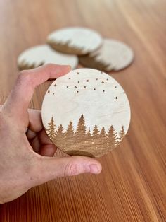 a hand holding a wooden ornament with trees and stars in the sky on it