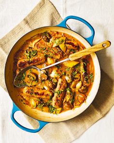 a blue pot filled with meat and vegetables on top of a white cloth next to a wooden spoon