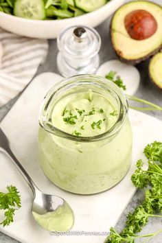 an avocado dip in a glass jar on a cutting board with spoons next to it