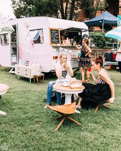 three women sitting at a picnic table in front of an old fashioned camper trailer