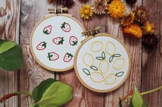 two embroidery hoops with fruit designs on them sitting next to some leaves and flowers