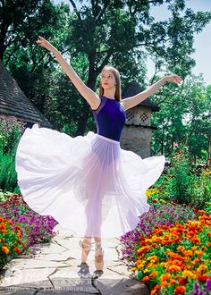 a woman in a purple top and white skirt standing on a stone path surrounded by flowers