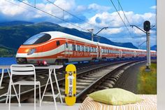 an orange and white train traveling down tracks next to the ocean with mountains in the background