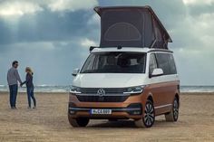 two people standing next to an orange and white vw van on the beach with its roof open