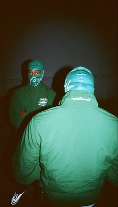 two men in green jackets standing next to each other with their faces covered by masks