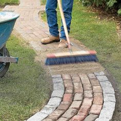 a man with a broom is cleaning the grass near a wheelbarrow and brick walkway