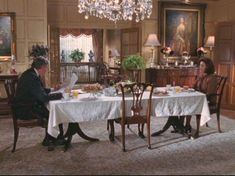 two people sitting at a dining table in a large room with chandelier and paintings on the walls