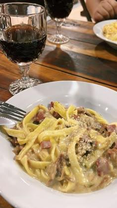 a white plate topped with pasta and meat next to a glass of wine on a wooden table