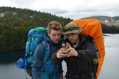 two men with backpacks looking at a cell phone in front of a body of water