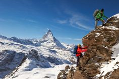 two people climbing up the side of a snow covered mountain with mountains in the background